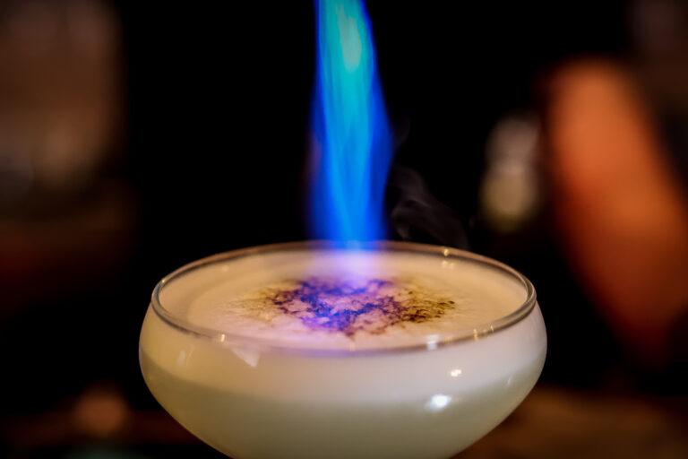 Craft cocktail being finished with a torch flame behind a bar